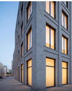 Corner of a tall brick building with large illuminated windows along a quiet street at dusk, sidewalk in foreground.