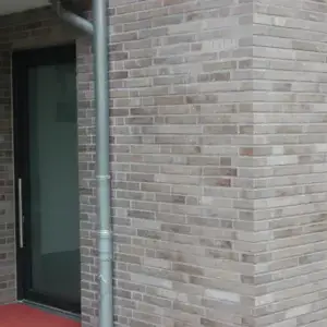 Brick exterior corner featuring a gray metal downspout beside a glass entrance door and a red entry mat outside.