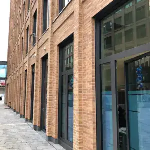 Brick building with tall glass storefront windows along a paved sidewalk; reflections of nearby structures in the glass doors.