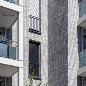 Modern apartment building exterior with gray brick, white frames, glass balconies, and string lights above.