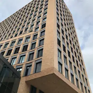 Low-angle view of a tall tan brick office building with a grid of dark-framed windows against a cloudy sky; blue balloons in the lower left corner.